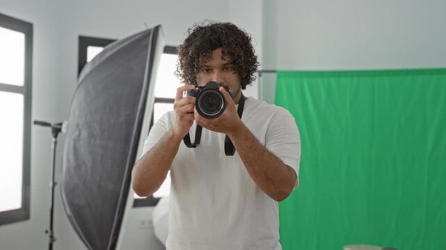 Man photographer in white tshirt holding camera to eye with strap, softbox and green screen in studio; focused creativity.