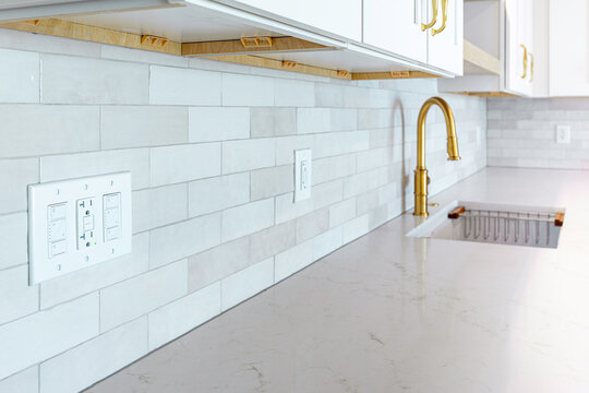 Close-up view of a three-gang electrical wall plate with integrated light switches and a GFCI outlet installed on a white tile kitchen backsplash near a brass faucet and quartz countertop, USA