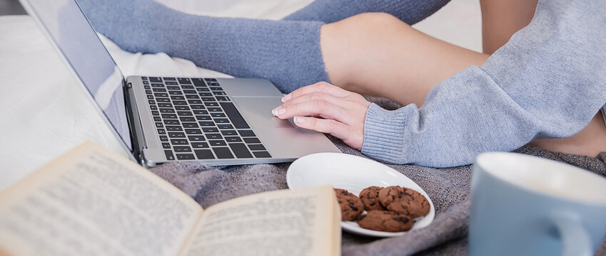 Woman in sweater with book and computer with cup of hot chocolate cookies winter time at home comfortable bedroom scene with laptop open book on bed, creating cozy remote work