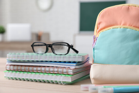 Stack of different notebooks with eyeglasses and backpack on wooden desk in classroom