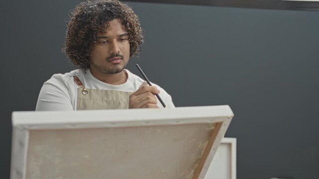 Man inspects paintbrush with raised hand while peering at canvas on easel in art studio; creative contemplation.