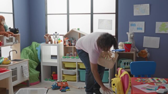Man picks up basket and bends to pick toys, hand holding wicker basket visible among colorful toys, shelves and play tent in kindergarten building; patience.
