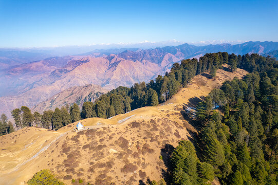 Aerial view of the Chakrata hill station featuring a small temple on a mountain ridge surrounded by pine forests and distant Himalayan peaks in Chakrata, Uttarakhand, India.