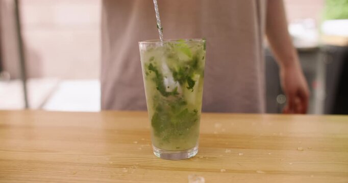 Stirring mojito. Refreshing drink. Unrecognizable bartender hand preparing cocktail in glass with crushed ice natural mint leaves lime at bar counter.