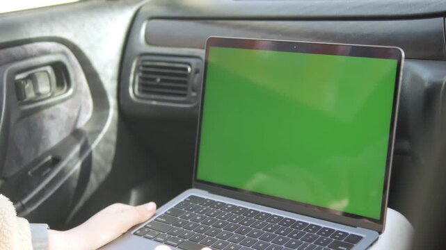 Laptop with green screen sits on car seat. Hand opens lid, then types on keyboard. Car interior shows dashboard and door controls. Sunlight enters from window. No animals or plants visible