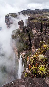 A breathtaking aerial view of Mount Roraima tepui flat-top mountain surrounded by clouds, featuring dramatic waterfalls cascading down its cliffs and a beautiful rainbow in the sky.