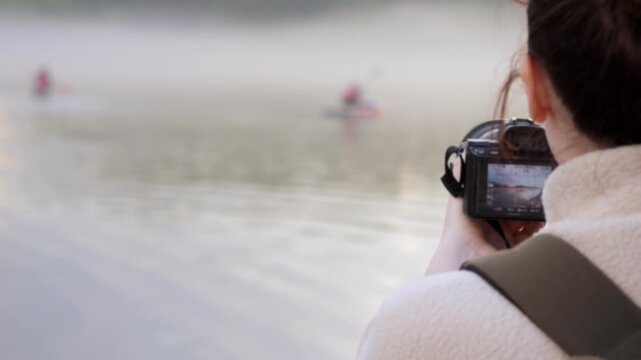 Photographer captures misty lake with rowers in early morning. Fog blankets water, softening distant trees. Person holds camera steady, focusing on boaters. Calm scene highlights natural beauty