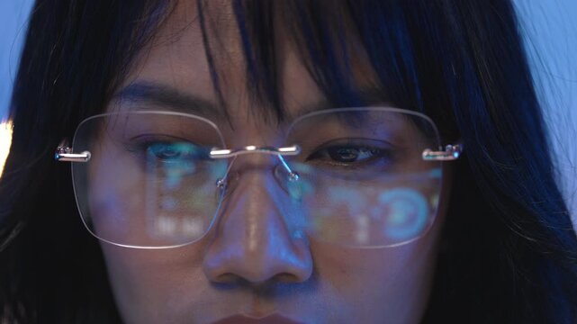 Macro close up of rimless eyeglasses on Asian woman face with bright digital programming code lines reflecting on the transparent lenses inside a dark blue office