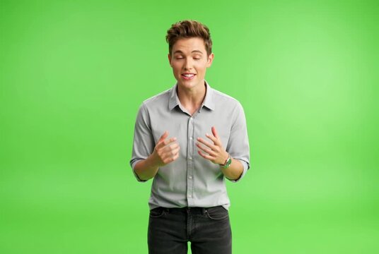 A friendly enthusiastic young Caucasian man in a casual grey shirt and jeans stands confidently smiling happily waving his open hand in a welcoming greeting gesture looking directly into the camera ch
