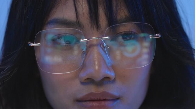 Portrait of a serious young Asian female software engineer wearing transparent rimless glasses concentrating on lines of code displayed on her monitor screen at night