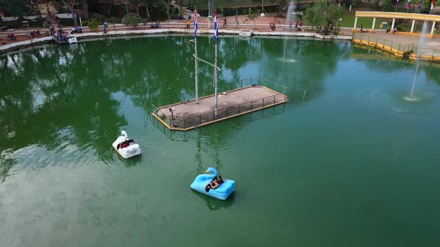 Drone aerial footage of blue and white swan paddle boats floating around the central platform at Ykua Pyt&atilde; lagoon surrounded by park walkways, fountains, and visitors near Villarrica, Paraguay.