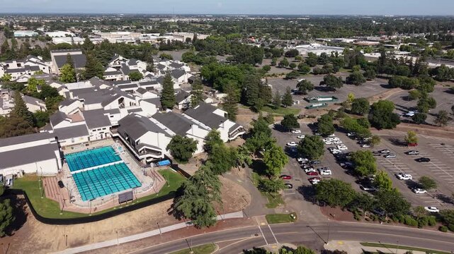 High-angle drone footage orbiting the swimming pool at the San Joaquin Delta College campus in Stockton, California, showing the surrounding buildings, parking lot, and cityscape on a sunny day