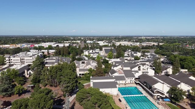 Sunny aerial perspective of the San Joaquin Delta College campus, featuring academic buildings, lush green trees, a large swimming pool, and the surrounding cityscape of Stockton, California