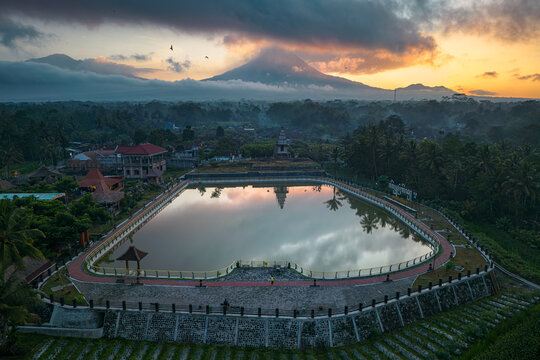 Aerial view of Embung Mranggen reservoir reflecting the sky with Mount Merapi volcano under a dramatic sunrise in Magelang, Central Java, Indonesia.
