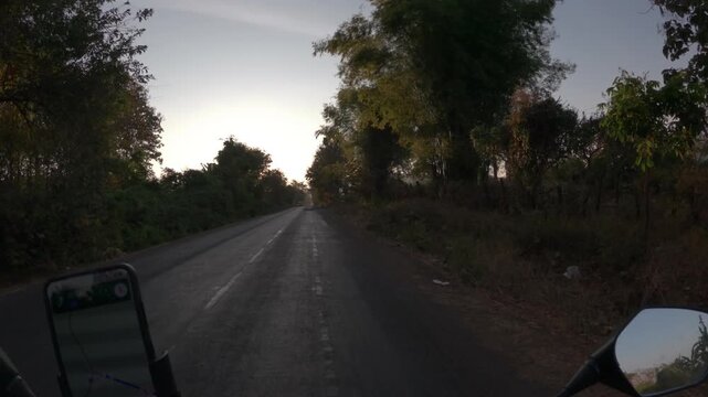 POV shot riding a motorbike in the early morning as the rising sun shines through gaps in the tree branches that line the road.