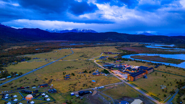 Aerial view of Rio Serrano Hotel Spa with illuminated buildings at twilight, a winding river, and snow-capped mountains in Torres de Paine, Magallanes, Chile.