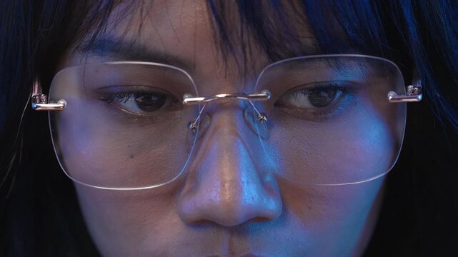 Extreme close up frontal portrait of a young Asian female programmer wearing modern transparent rimless eyeglasses with subtle blue light reflecting on her face