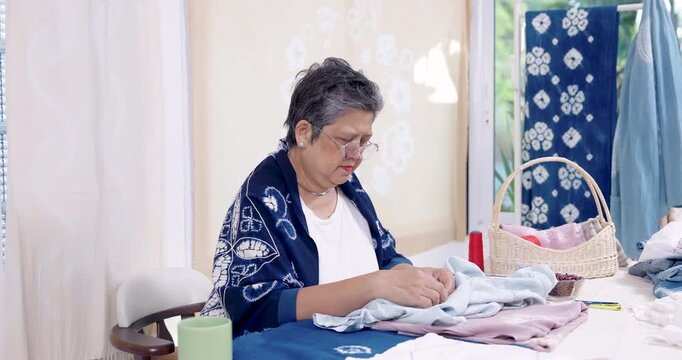 Senior Asian woman artisan hand stitches denim slowly, pulls red thread, tightens resist line for shibori pattern, calm home studio tutorial scene, slow craft process for small business order.