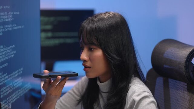 Young Asian woman software developer holding a smartphone close to her mouth giving voice commands while sitting in front of monitors filled with programming code