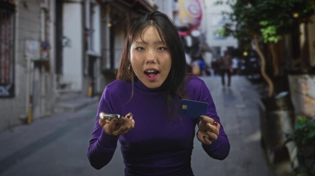 Young chinese woman holding smartphone and creditcard, mouth open and looking up on street; surprise discovery.