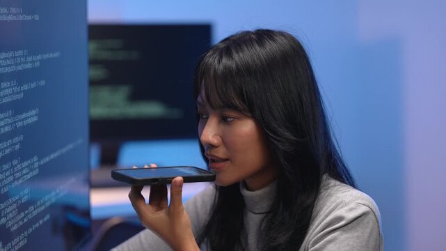 Young Asian female software engineer in turtleneck sweater using voice command on her smartphone while working late at the office with bright screens of code behind her