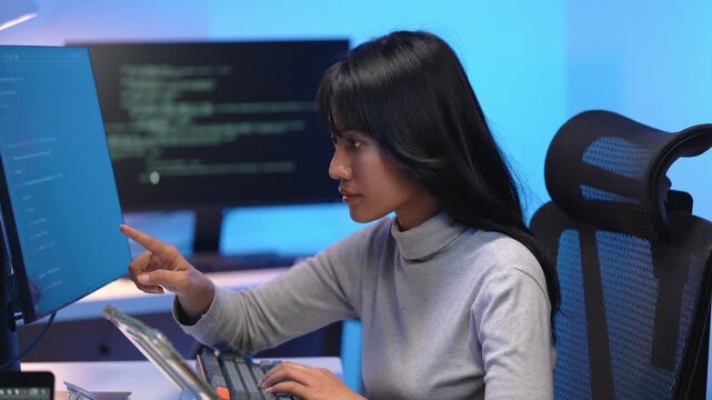 Young Asian woman software developer in a casual sweater typing on a laptop keyboard at her desk with dual monitors displaying programming code in a dark blue office