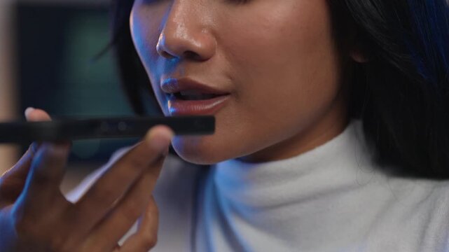 Extreme close up of a young Asian woman in a white turtleneck sweater holding her smartphone near her mouth using voice recognition or voice command technology at night