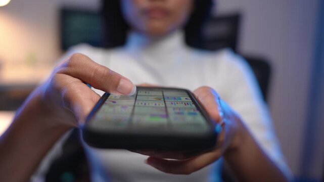 Close up of young Asian woman hands holding a smartphone displaying a colorful application interface with soft bokeh background of a dark modern office at night