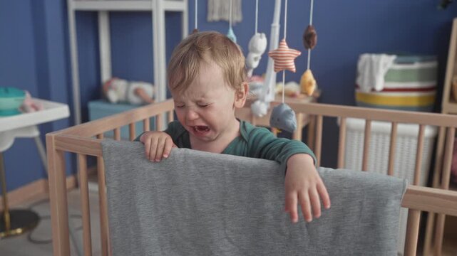 Boy crying with hands gripping crib rail and gray blanket over rail in building nursery, mobile toys above; distress need comfort.