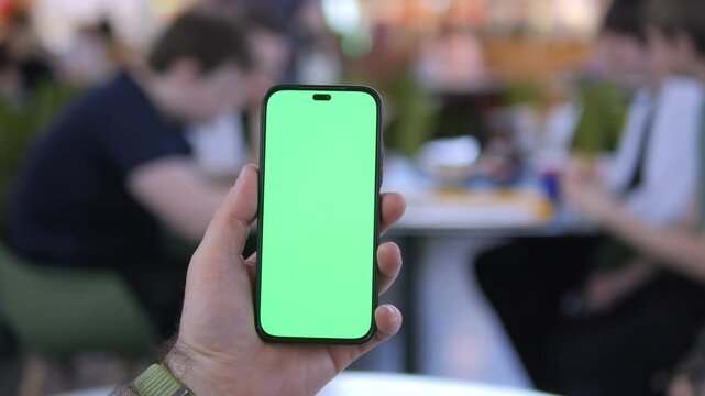 Hand holds phone with green screen. Background shows blurred cafe scene. People sit at tables, eating or drinking. Warm lighting from outdoor seating area. Focus stays on phone throughout clip