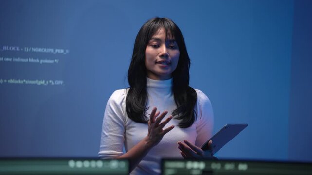 Young Asian woman software engineer holding a digital tablet while presenting and teaching a programming class in front of a large bright screen displaying source code