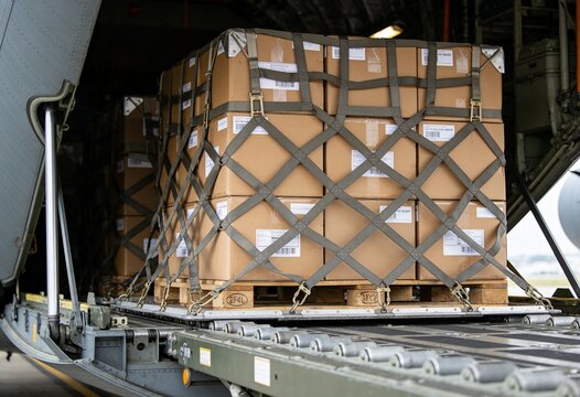 A large cargo pallet loaded with numerous cardboard boxes, secured by a cargo net, positioned within a transport aircraft's bay.