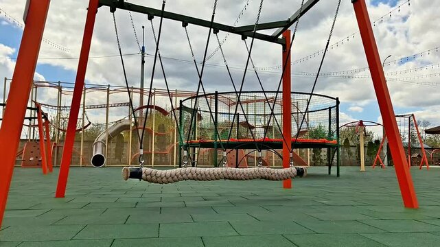 Large circular rope swing with red metal supports in a modern playground, surrounded by trampolines and equipment, set on green safety tiles under a partly cloudy sky