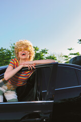 Fototapeta premium Joyful woman leans from a car window, laughing with a retro hairstyle and striped shirt, bright outdoor scene, cinematic color, casual lifestyle moment, playful mood.