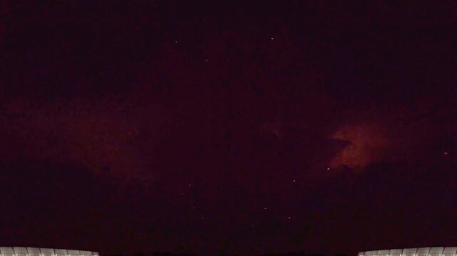 Dramatic night sky lightning storm flashing through dark storm clouds over a stadium roof structure.
