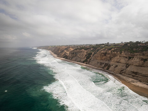 Aerial view of the rugged cliffs and breaking waves along the sandy coastline under a cloudy sky in San Diego, California, United States.