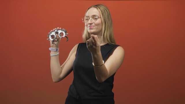 Woman holding optometry trial frame and beckoning with hand while smiling and wearing glasses in studio; friendly confidence.