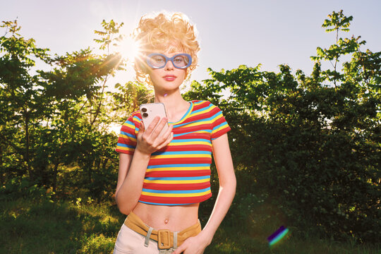 Young woman with curly blonde hair and blue sunglasses wears a colorful striped crop top in a sunny park while looking at her smartphone exuding playful retro vibes