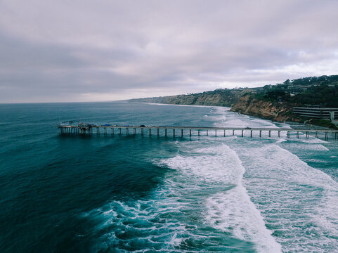 Aerial view of Scripps Pier stretching into the Pacific Ocean with crashing waves and coastal cliffs under a cloudy sky in San Diego, California, United States.