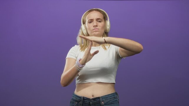 Blonde woman wearing headphones makes timeout t gesture showing midriff and wristband in studio; calm contemplation.