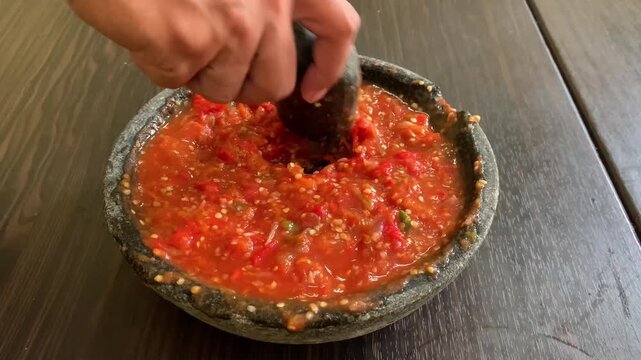 Close-up of a person crushing fresh red chili sambal using a traditional stone mortar and pestle (cobek) on a wooden table. Authentic Indonesian spicy condiment preparation.