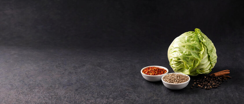 Head of green cabbage and spices: red pepper flakes, cumin, black peppercorns, and cinnamon sticks on a dark stone background.
