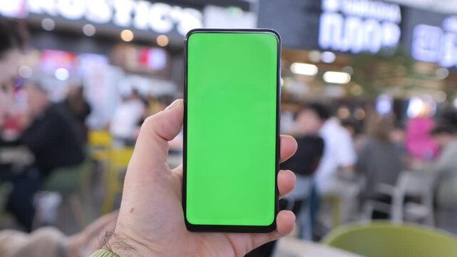Hand holds phone with green screen. Background shows blurred indoor market area. People sit and walk behind. Hand steady, no movement. Focus on phone screen only. Background not detailed