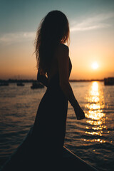 Fototapeta premium Woman silhouette standing on boat during golden hour sunset, Female traveler on water yacht at sunrise with elegant dress