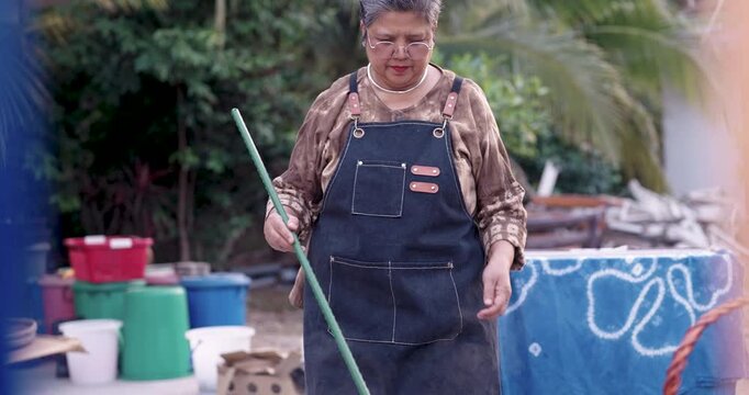 Senior Asian craftswoman stands near dye station holding long pole, preparing next indigo dye step for shibori fabric, outdoor workshop equipment ready for sustainable heritage textile business.