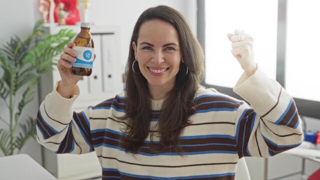 Woman holding a medicine bottle, smiling and raising a fist in a clinic building with binders and an anatomy model on a desk visible; relief recovery.