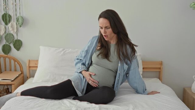 Pregnant woman clutching belly while seated on white bed in bedroom, bracing and grimacing during contractions; labor pain resilience.