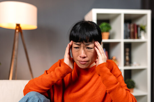 Asian woman lying on sofa with severe menstrual cramps. Sick female patient holding painful stomach with hands, suffering from abdominal pain, digestive problem. Healthcare and medicine snapshot.