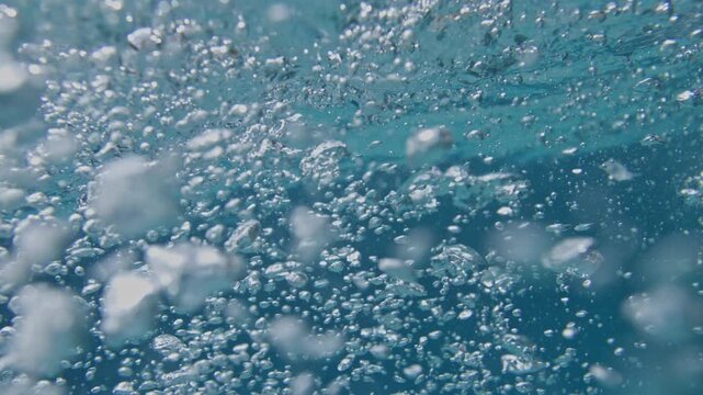 Thousands of suspended underwater bubbles rise through turbulent tropical Atlantic water while refracted sunlight creates abstract blue marine textures beneath the surface in slow motion