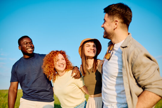 Group of diverse young adult friends smiling and laughing together outdoors under a clear blue sky, embracing happiness